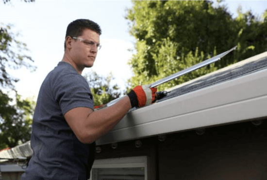 A person wearing safety glasses and gloves is cleaning debris from a house gutter with a tool, standing on a ladder near the roof. The scene highlights the importance of safety, with trees in the background and gutter guard manufacturer innovation in mind.
